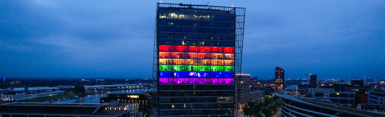 Bild des Weser Towers vor dunklem Abend-Himmel. Der Weser-Tower hat mittig sechs Büroetagen in unterschiedlichen Farben beleuchtet, sodass eine Regenbogenflagge entsteht. 