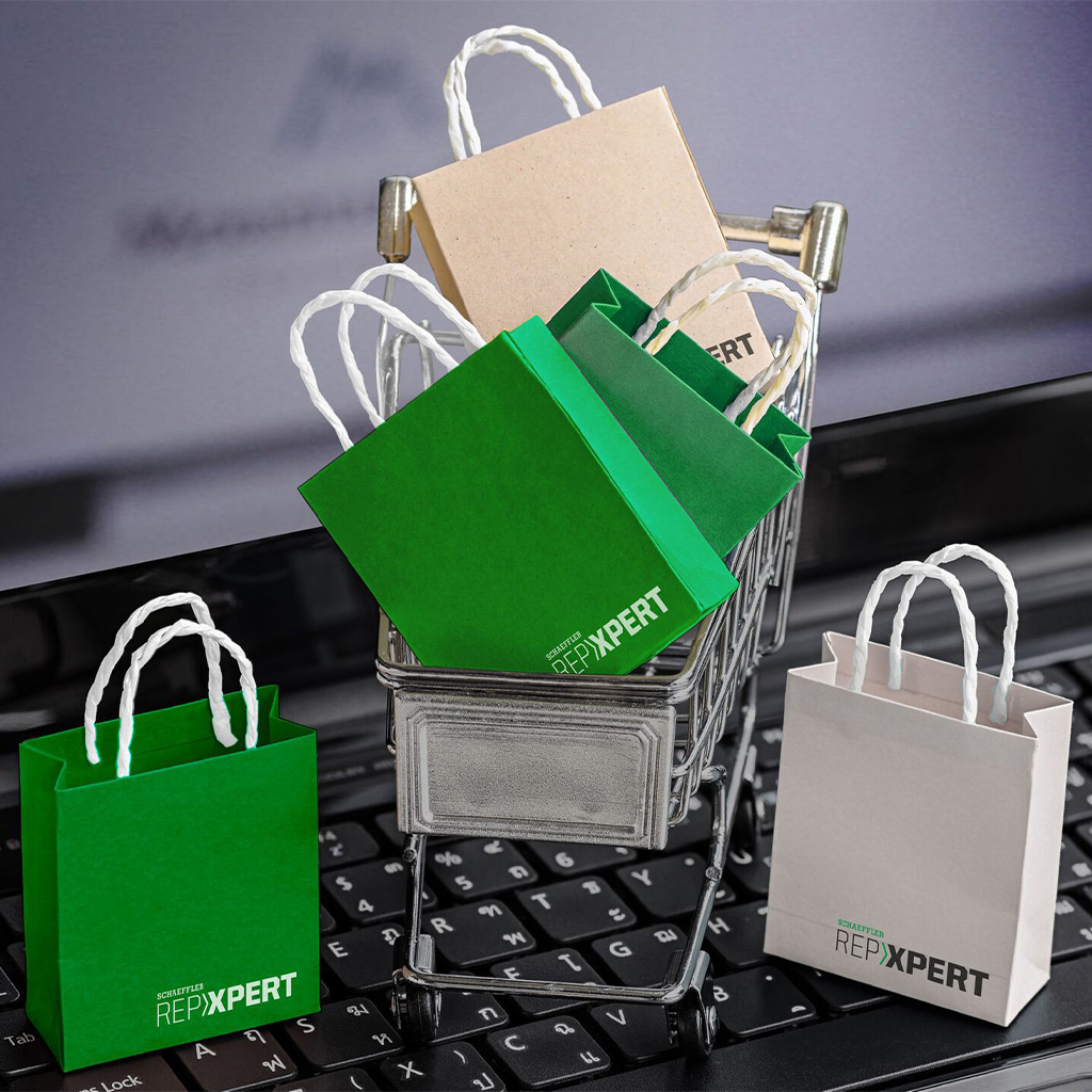 Ein Laptop füllt das ganze Bild, wobei auf der Tastatur ein kleiner Einkaufswagen steht, der mit grünen und weißen kleinen Tüten gefüllt ist. Rechts neben dem Einkaufswagen steht eine weiße Tüte, links daneben eine grüne. Die Einkaufstüten tragen den Schriftzug "REPXPERT" an der unteren Kante.