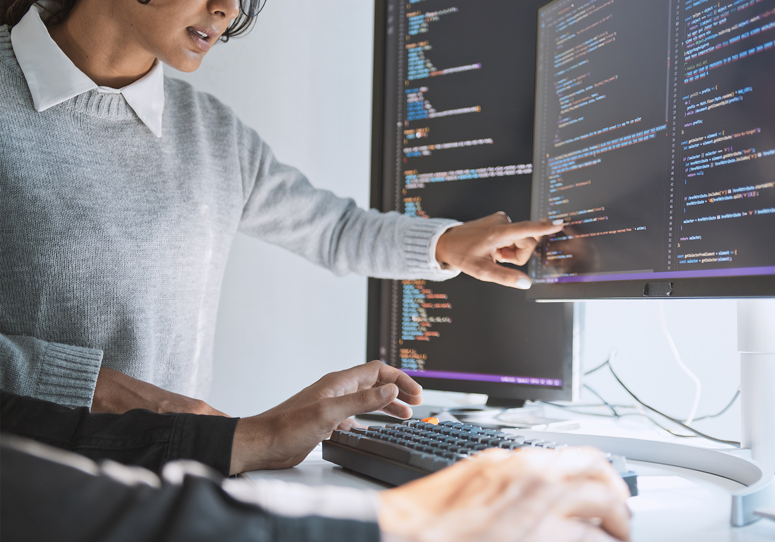 Foto eines Bildschirms auf dem ein Code abgebildet ist. Davor Hände auf einer Tastatur und eine Frau, ab dem Mund abgeschnitten, die auf den Bildschirm zeigt